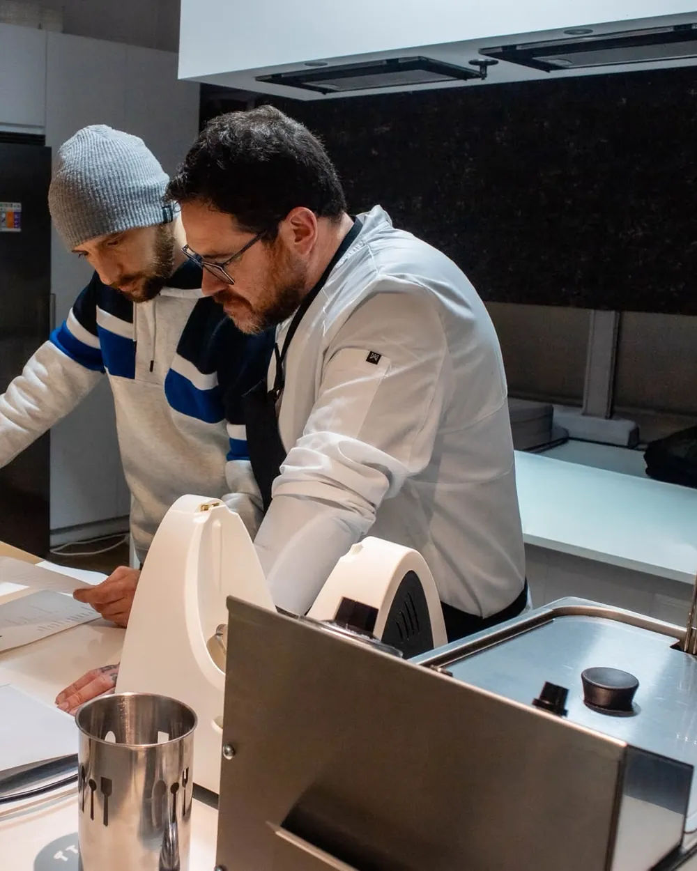 El cocinero, con chaquetilla blanca, y dos compañeros revisan unos papeles en la encimera de la cocina vista